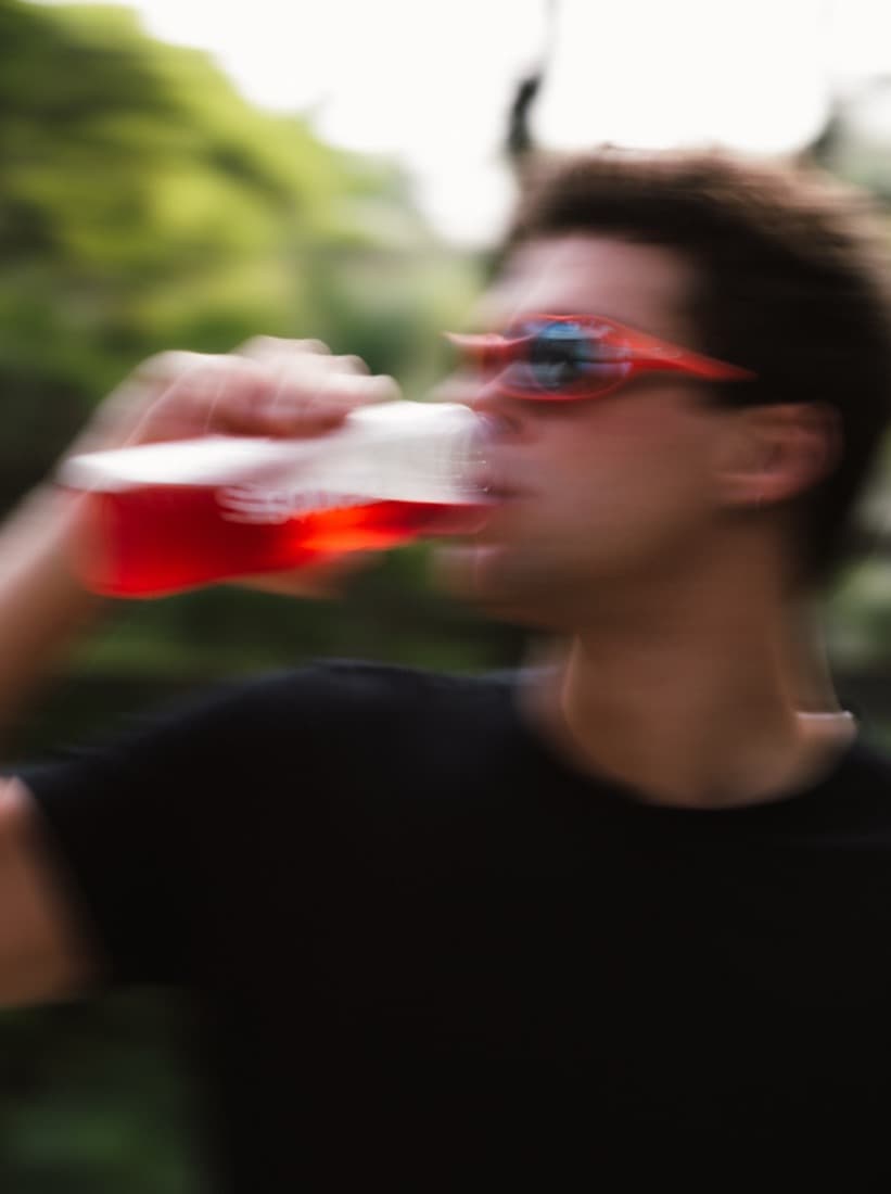 Homem de óculos escuros vermelhos e camiseta preta bebe Squezzy direto da garrafa ao ar livre, numa foto com borrão de movimento.