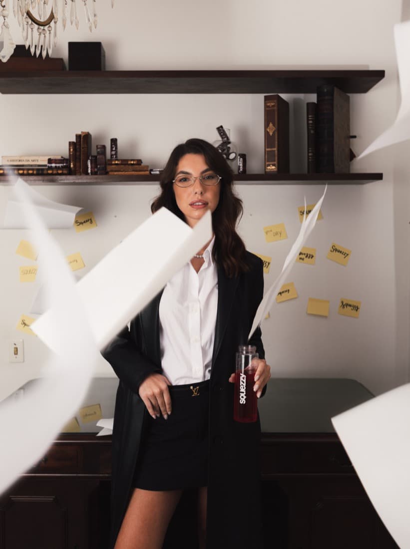 Mulher de óculos, camisa branca e blazer preto segura uma garrafa com Squezzy diante de uma estante, com folhas de papel voando à frente.